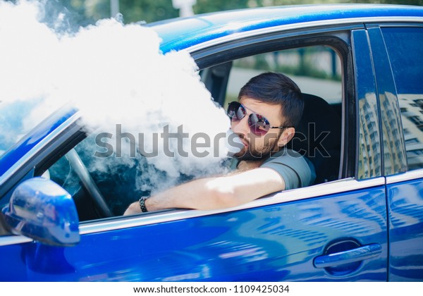 A guy with a beard in sunglasses is\
sitting in the car and smokes an electronic\
cigarette