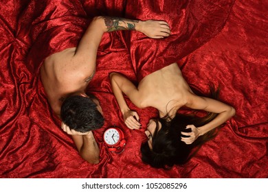 Guy with beard and tattoos touches pretty ladys back in bed, top view. Man and woman with half covered bodies lie near red clock. Couple in love on red sheets. Love, sex and early morning concept