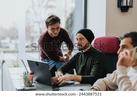 A guy in a black cap commits an error in the project tasks, working hard to rectify it, while his male colleague sitting next to him playfully mocks the situation.