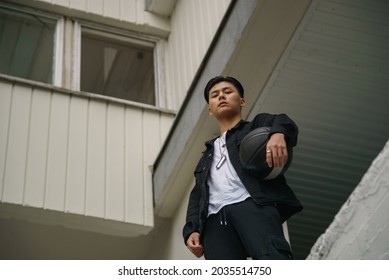 Guy In Black Holding A Basketball. Young Man With Black Ball Chewing Gum While Standing Against Multi-storey Building