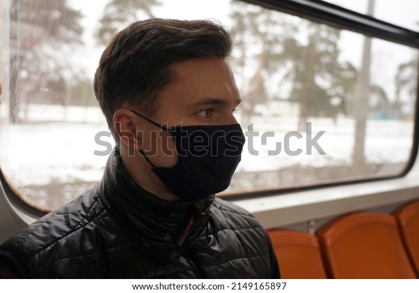 A guy in a black mask sits in a subway car near\
the window