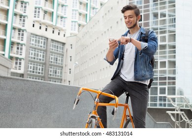 The guy in a blue denim jacket standing on city background. a young man near orange bicycle. Smiling student with bag looking at phone - Powered by Shutterstock