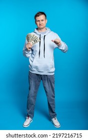 A Guy In A Blue Sweatshirt And Jeans Holds A Fan Of Dollars And Points His Finger At Himself, Frowning. Earned Little. Full Height. Blue Background