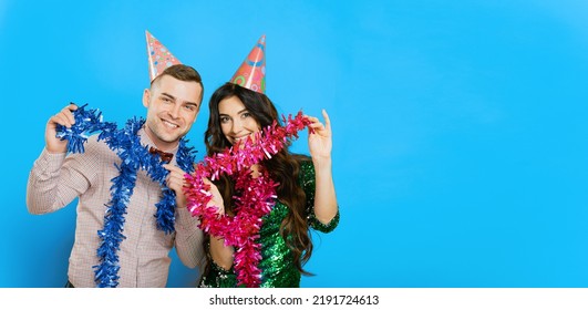A guy in a bow tie with a girl 20-25s in party hats pose with New Year's tinsel in their hands and look at the camera on a blue isolated background. Party banner with copy space
