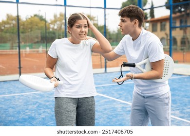 Guy consoling frustrated young female teammate after losing paddle tennis match while standing on open court on warm autumn day. Agony of defeat and friendly support - Powered by Shutterstock