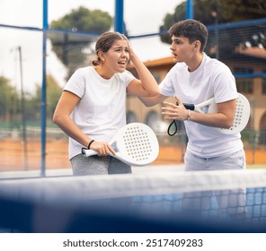 Guy consoling frustrated young female teammate after losing paddle tennis match while standing on open court on warm autumn day. Agony of defeat and friendly support - Powered by Shutterstock