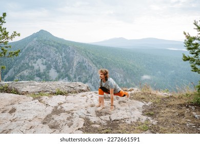 The guy does stretching of muscles and ligaments standing in nature in the mountains, yoga practice on a rock, lunge in front, fitness in the morning with a beautiful view, workout. - Shutterstock ID 2272661591