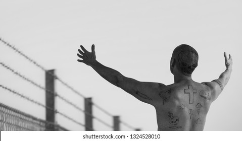 Guy With Doodle Tattoos With Arms Reached Out At Barbed Wire Fence. Criminal, Convict Riot And Prison Tattoo Concept. Image Montage.