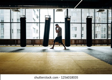 Guy Dressed In The Grey T-shirt And Black Shorts Stands On Guard And Works Out A Boxing Punch Next To Hanging Punching Bag Against The Background Of Panoramic Windows In The Gym