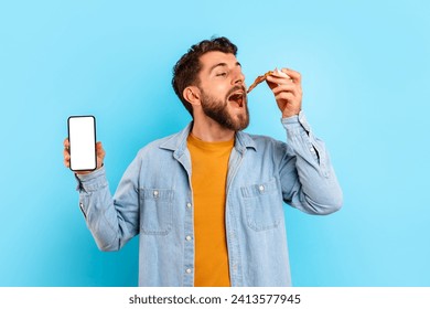 Guy eats pizza and shows mobile phone with empty screen over blue studio backdrop. Hungry man savoring tasty unhealthy junk food and advertising application for pizzeria delivery. Mockup