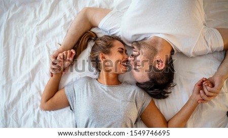 Guy and a girl in a cozy home environment. Happy man and woman lying in the bedroom stock photo. Top view of smiling young couple cuddling in bed in morning.  Beautiful pair of lovers hug and kiss