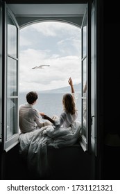 A guy and a girl in light shirts are sitting wrapped in a blanket on the windowsill of a large open window with beautiful views of the sea and mountains. A seagull flies by. Cozy atmosphere. Lovers