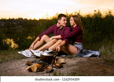 a guy and a girl in red checkered shirts are sitting by the river and the guy is straightening a branch with a bonfire - Shutterstock ID 1162954309