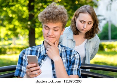 A guy with a girl in the summer in nature. A young man looking into the phone reading a correspondence. The girl spies from behind. The concept of mistrust. Peeping at the interlocutor.