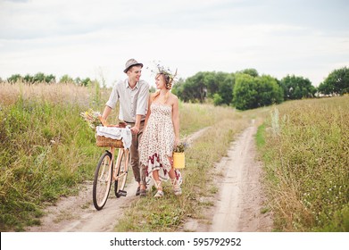A guy and a girl are walking along a trail in the field smiling at each other. They are dressed in rustic style. In his hands, a bicycle with a basket and food. Honeymoon. - Powered by Shutterstock