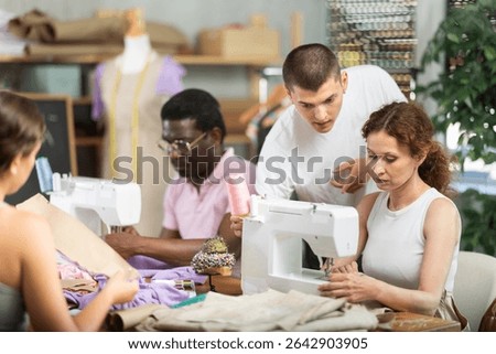 Guy head of school of applied professions helps student to sew product, other participants work with materials and patterns, communicate and spend time in company of like-minded people..