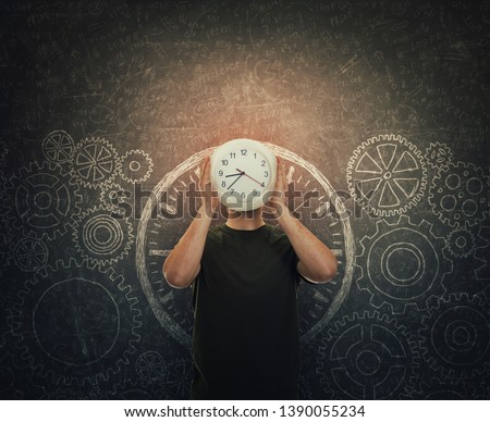 Guy hiding face holding a clock instead of head stands over dark blackboard with drawn gears and cogwheels. Business time management concept. People goals, plan and take action. Schedule efficiency.