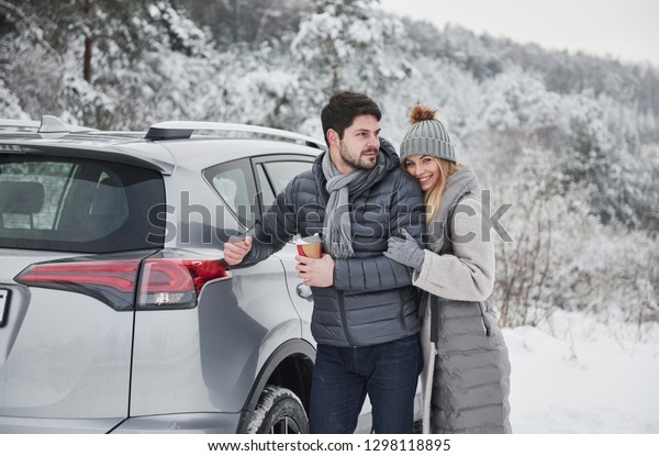 Guy holding cup with warm drink and leaned on his\
car while girl hugs him. Nice couple have weekend at countryside at\
winter time.