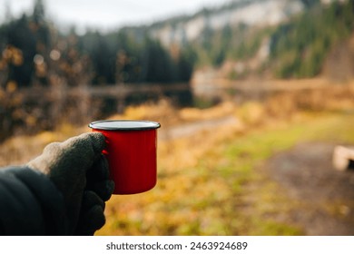 A guy holds a red mug on the riverbank in autumn - Powered by Shutterstock