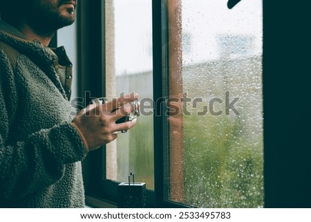A guy at home holding a cup of coffee looking outside the window on a rainy day