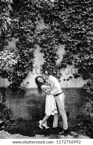 The guy kisses his girlfriend in a cheek against the background of the wall and plants with leaves on a black and white photo