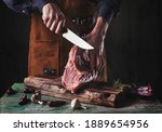 A guy in a leather apron is slicing raw meat. The butcher cuts the pork ribs. Meat with bone on a wooden cutting board.