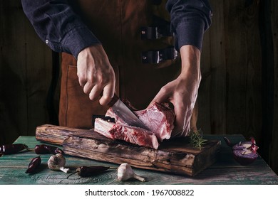 A guy in a leather apron is slicing raw meat. The butcher cuts the pork ribs. Meat with bone on a wooden cutting board. - Powered by Shutterstock