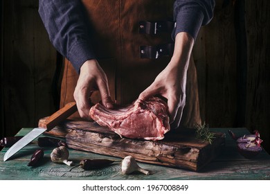 A guy in a leather apron is slicing raw meat. The butcher cuts the pork ribs. Meat with bone on a wooden cutting board. - Powered by Shutterstock