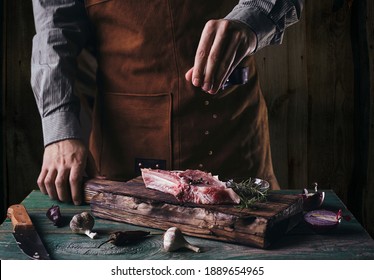 A guy in a leather apron sprinkles black pepper on a steak with a bone. Raw meat on a wooden cutting board.