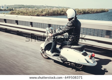 Guy in leather jacket and helmet is riding on scooter through the city bridge
