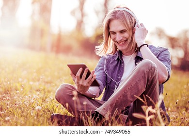 Guy looking something on the tablet and listening to music,he is sitting on the grass in summer warm park. - Shutterstock ID 749349055