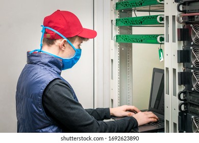 A Guy In Medical Mask Is Working In Front Of A Laptop. Programmer In Virus Protection Tools Works In The Server Room Datacenter. Maintenance Of Computer Equipment Under Quarantine.