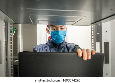 A Guy In Medical Mask Is Working In Front Of A Laptop.Programmer In Virus Protection Tools Works In The Server Room Datacenter. Maintenance Of Computer Equipment Under Quarantine.