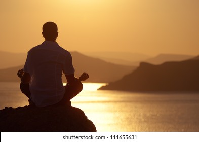 Guy meditating at sunset sitting on a rock by the sea