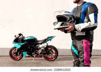 guy motorcyclist in professional protective equipment stands near sports motorcycle and holds helmet against white wall, professional motorcyclist with bike on the street