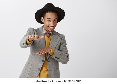 Guy is on same wave with us. Portrait of flirty confident male model in fashionable outfit with sense of style, wearing hat and making gestures with palms towards camera, smiling broadly