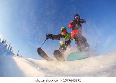 A Guy On Skis And A Girl On A Snowboard Perform A Trick Against The Background Of A Blue Winter Sky.