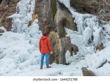 a guy in an orange winter jacket is walking and drinking a hot drink near a frozen waterfall, a concept of adventure, new discoveries, freedom and travel