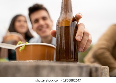 Guy Pick Up Beer From The Table Outdoors. Closeup On The Bottle With No Label. People Eating Take Away Food Outdoors And Having Fun Drinking Beers Together. Blurred People Faces On The Background.
