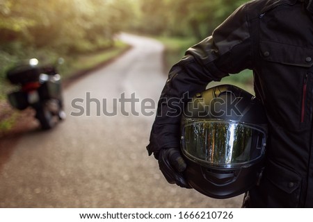 Guy in raiders clothes, gearing up. A young men stands near an motorcycle in nature during sunset. Biker with motorcycle on the road. Motorcycle and safe driving. Helmet and motorcycle.