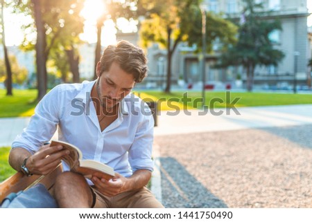 Guy reading book with text space. Young man reading book in park copy space. Man sitting on bench reading book in town. Happy positive guy reading book