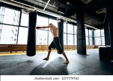 Guy In Red Boxing Gloves With A Naked Torso Dressed In The Black Shorts Hits Punching Bag  In The Gym  Against The Background Of Panoramic Windows