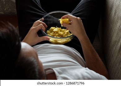 The Guy In The Shirt Is Lying On The Couch, Eating Chips And Watching A Sports Channel. The Concept Of Laziness, Frustration, Procrastination, The Person At Home.