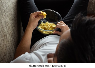 The Guy In The Shirt Is Lying On The Couch, Eating Chips And Watching A Sports Channel. The Concept Of Laziness, Frustration, Procrastination, The Person At Home.