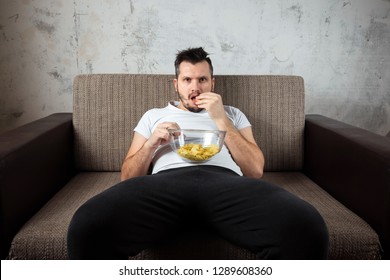 The Guy In The Shirt Is Lying On The Couch, Eating Chips And Watching A Sports Channel. The Concept Of Laziness, Frustration, Procrastination, The Person At Home.