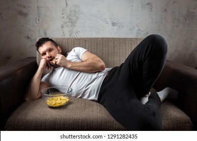 The guy in the shirt is lying on the couch, eating chips and watching a sports channel. The concept of laziness, frustration, procrastination, the person at home.