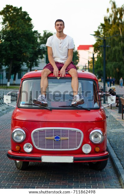 The guy sits on the roof of his retro car, in the\
middle of a beautiful\
street.