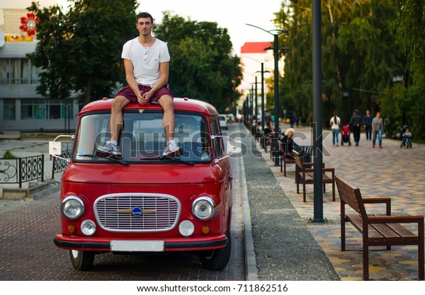 The guy sits on the roof of his retro car, in the\
middle of a beautiful\
street.