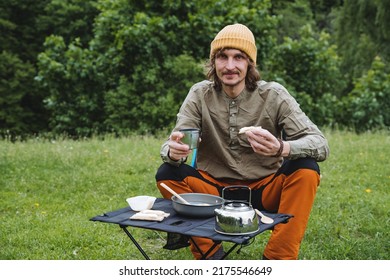 A guy sitting in nature smiling holding a mug with tea and a sandwich with butter, breakfast on a hike, a vacation in the woods, a hipster relaxing, hiking. High quality photo