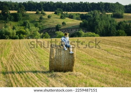 A guy sitting on a large bale of straw in an empty field of cut wheat